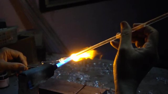 The man's hand is holding the glass stick to the fire to melt and mold, craft art from the blown glass.