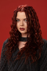 Beautiful sexy woman is posing in a knitted dress on a red background. Evening makeup. Long, healthy, red curly hair.
