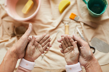 Above view closeup of adult woman gently holing hands of little girl in pottery workshop for family, copy space