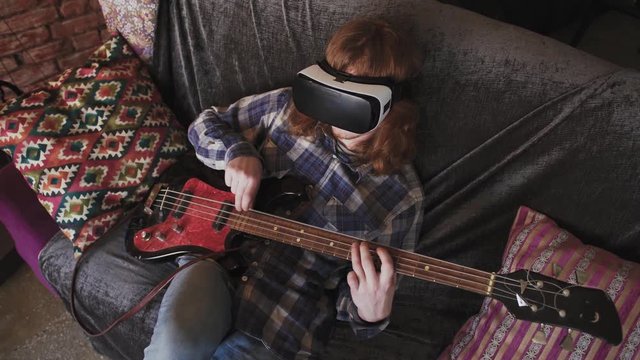 Young redhead man musician playing bass guitar in VR glasses at home, sitting on sofa, slow motion