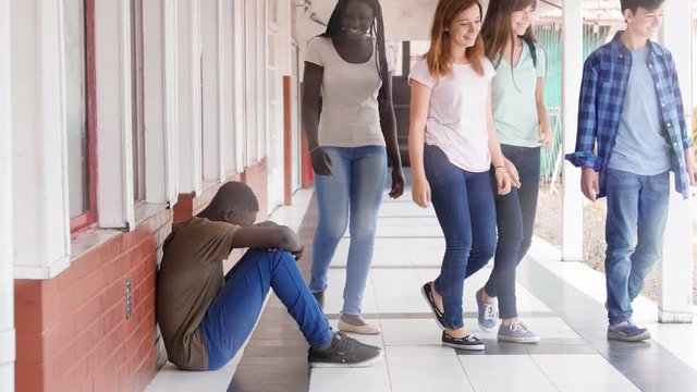 Afroamerican teenager boy seated on school hallway, bullying concept with schoolmates having fun of him