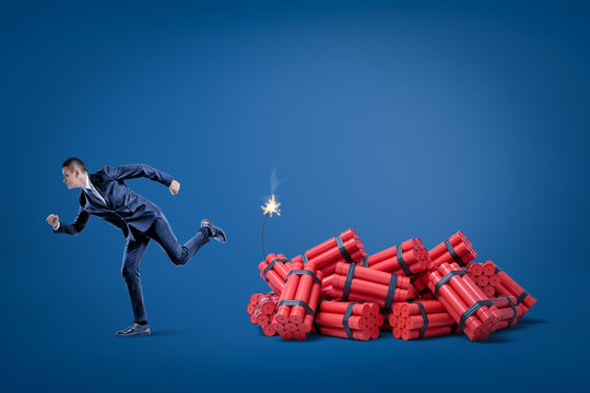 Businessman Running Away From Pack Of Red Tnt Dynamite Sticks With Lighted Fuse On Blue Background