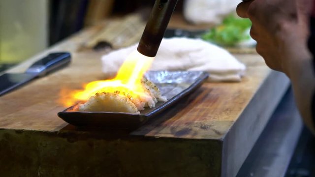 Slow Motion of asian cheff burns a slice of raw fish with blowtorch for make sushi caramelize. Man cooking in japanese restaurant. Searing and burning fresh sashimi on a kitchen background, Japan -Dan