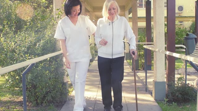 Nurse helping elderly woman walking with stick, front view with sunlight coming from outside