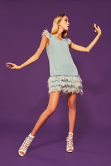 Image of pretty young blonde girl in blue cocktail dress dancing on purple background in studio