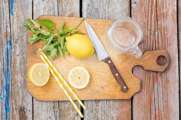 Chopping cutting board, sliced lemon, jar, mint leaves, knife and straw on wooden table. Cocktail preparation. Summer. Top view.