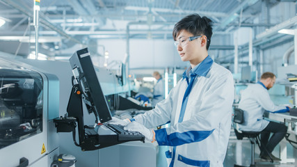 On High Tech Factory Asian Engineer Uses Computer for Programing Pick and Place Electronic Machinery for Printed Circuit Board Surface Mount Assembly Line. Production of PCB with SMT Machinery.