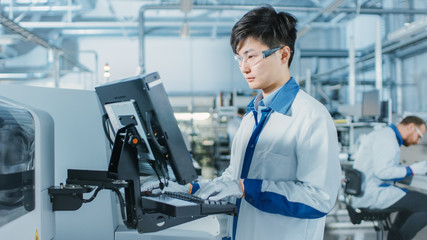 Fototapeta premium On High Tech Factory Asian Engineer Uses Computer for Programing Pick and Place Electronic Machinery for Printed Circuit Board Surface Mount Assembly Line. Production of PCB with SMT Machinery.