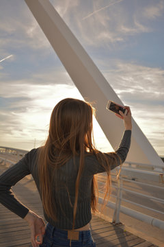 A beautiful and young blonde and Caucasian woman stands on a bridge to take a selfie on her mobile phone in a sunny day during the sunset