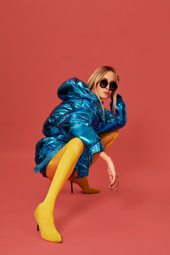 Young confident blonde girl in blue jacket squats in studio before camera, red background