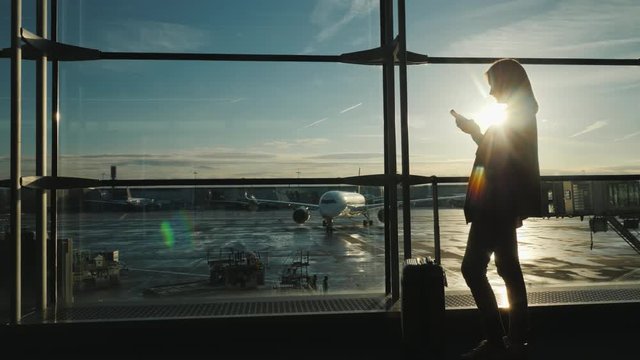 Passenger with a smartphone in the airport terminal. The silhouette illuminates the sun, behind the large windows you can see the airliner