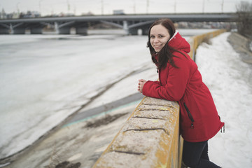 Beautiful girl in a red jacket on the pier in the spring posing against the water. Woman on the waterfront