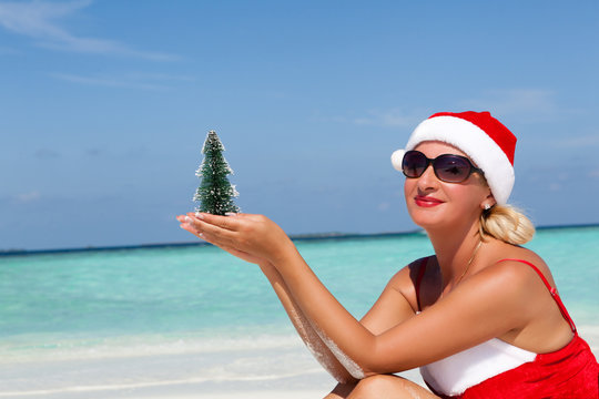 Santa woman sitting at the beautiful white sand beach and holding the Christmas tree. Copy space