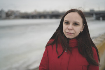 Beautiful girl in a red jacket on the pier in the spring posing against the water. Woman on the waterfront