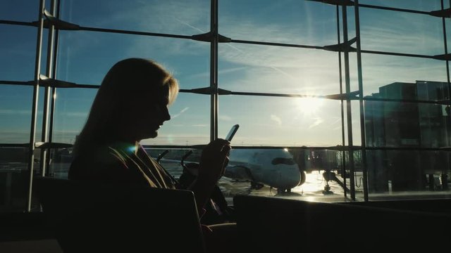 Silhouette of a woman, sitting by the window in the airport terminal, using a smartphone