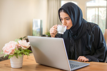 Young Arab woman drinking coffee and reading news on laptop at home