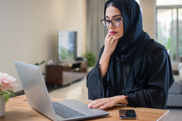 Pensive or worried Arab woman in abaya using laptop at home
