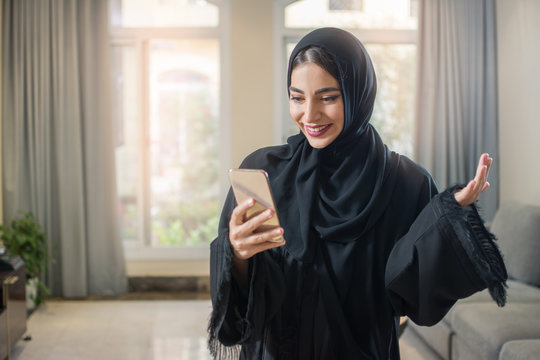 Portrait of cheerful muslim young woman in abaya using smartphone while standing in living room.