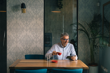 gray haired businessman using tablet and drinking coffee in restaurant