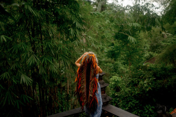 beautiful woman in long bright indian roach and blue boho clothes posing on the wood verande in tropical Jungle background in Bali
