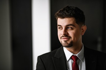 A handsome man in a black suit and a red tie posing at the big window. Business portrait of a man.