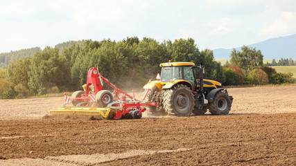 Yellow tractor pulls red sow mechanism over field, trees in background.