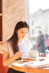 A young beautiful woman came to the cozy cafe in the morning to eat breakfast with a delicious granola sitting at a table near a large bright window