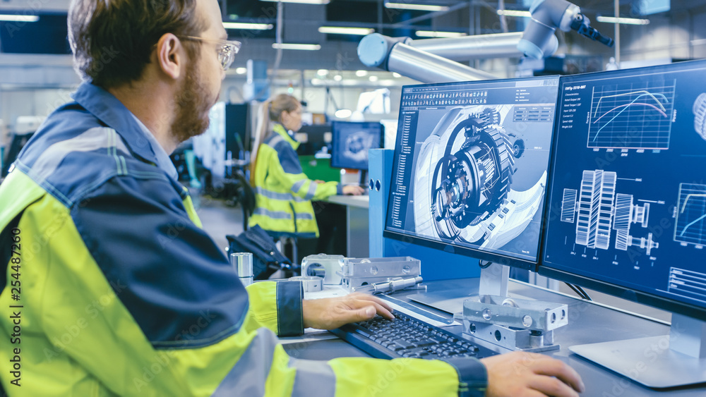 Fototapete At the Factory: Mechanical Engineer Works on Computer, Designs in CAD 3D Model of the Engine. In the Background Factory Workers and Manufacturing Process.