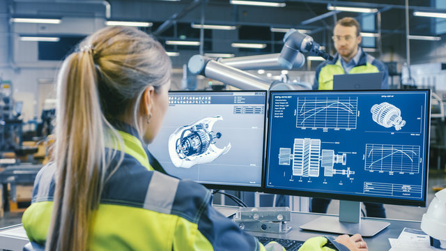 At the Factory: Female Mechanical Engineer Designs 3D Engine on Her Personal Computer. In the Background Male Automation Engineer who Uses Laptop for Programming Robotic Arm.