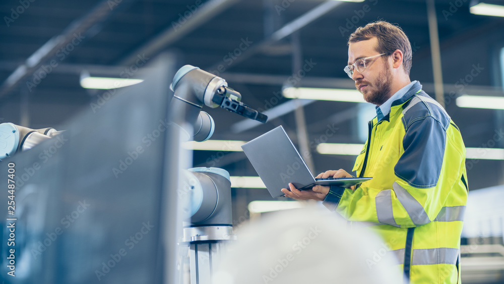 Poster At the Factory: Automation Engineer Uses Laptop for Programming Robotic Arm. New Era in Automatic Manufacturing Industry.