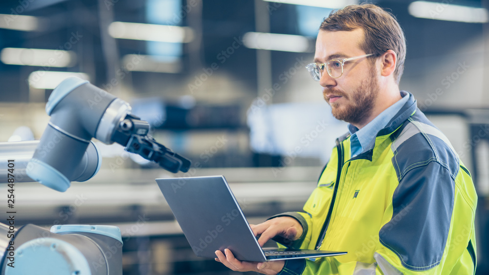 Poster At the Factory: Automation Engineer Uses Laptop for Programming Robotic Arm. New Era in Automatic Manufacturing Industry.
