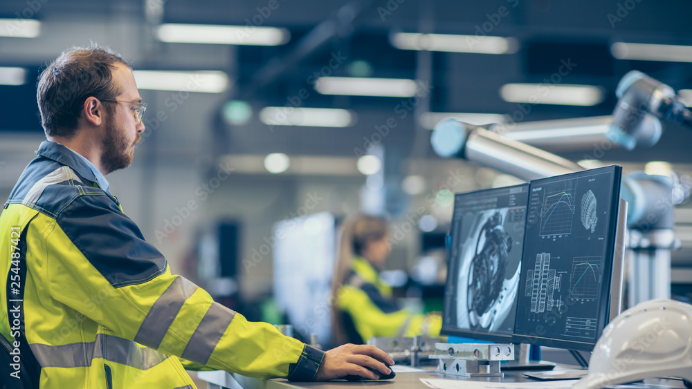 Leinwandbilder At the Factory: Mechanical Engineer Works on Computer, Designs in CAD 3D Model of the Engine. In the Background Factory Workers and Functioning Robotic Arm.