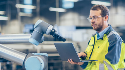 At the Factory: Automation Engineer Uses Laptop for Programming Robotic Arm. New Era in Automatic...