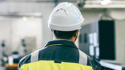 Back View Shot of the Industrial Engineer Wearing Protective Clothing Walks Through Modern Manufacturing Facility with Automatic Machinery Working in Background.