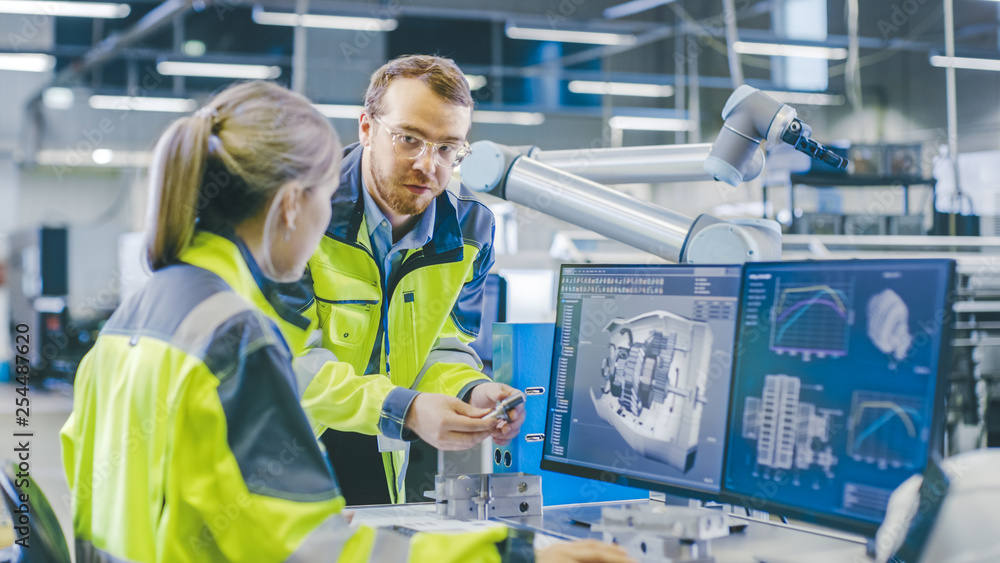 Poster At the Factory: Male Mechanical Engineer Holds Component and Female Chief Engineer Work on Personal Computer, They Discuss Details of the 3D Engine Model Design for Robotic Arm.