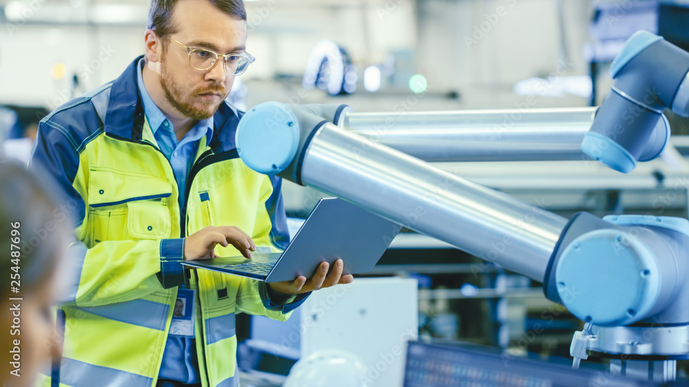 Fototapete At the Factory: Automation Engineer Uses Laptop for Programming Robotic Arm. New Era in Automatic Manufacturing Industry.