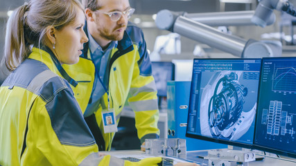At the Factory: Male Mechanical Engineer and Female Chief Engineer Work Together on the Personal Computer, They Discuss Details of the 3D Engine Model for Robotic Arm.