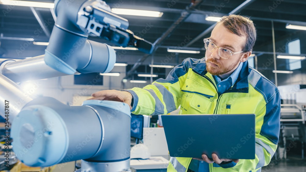 Poster At the Factory: Automation Engineer Uses Laptop for Programming Robotic Arm. New Era in Automatic Manufacturing Industry.