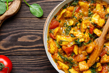 Top view on Bombay chicken curry with spinach, tomato and onion in a white frying pan on a kitchen wooden table. Indian traditional cuisine.