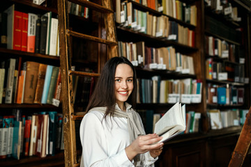 Young attractive student girl reading a book between library bookshelves