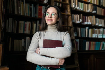 Brunette clever student standing near the bookshelves and holding big book at the library