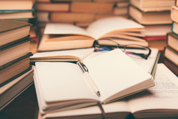 Education learning concept with open book and notebook on the table. Stack piles of books on reading desk and glasses in bookshelves background