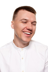 Charming handsome young man in a white shirt is making faces, while standing isolated on a white background