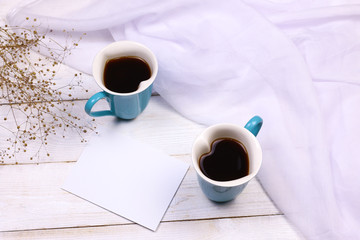 Heart shaped coffee cups with blank paper on white wooden background and transparent fabric. Love and romantic relationship concept mockup.