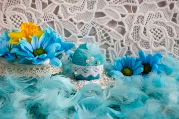 Wielkanocna kompozycja z niebieskimi pisankami, żółtymi i niebieskimi kwiatami oraz piórami na tle białej koronki © Monika