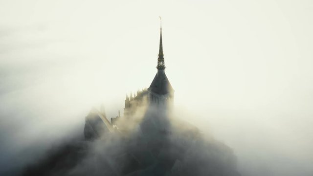 Aerial shot, fog clouds flowing over famous majestic Mont Saint Michel fortress castle during epic sunrise in Normandy.