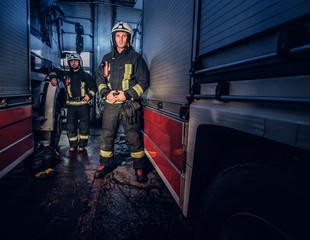 Fototapeta na wymiar Full-length portrait of two brave firemen in protective uniform standing between two fire engines in the garage of the fire dept