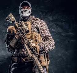 Private security service contractor wearing a balaclava skull and cap holding an assault. Studio photo against a dark textured wall - obrazy, fototapety, plakaty