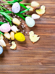 Flowers, painted Easter eggs and chickens on a brown wooden background with space for text. Easter concept