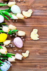 Flowers, painted Easter eggs and chickens on a brown wooden background with space for text. Easter concept
